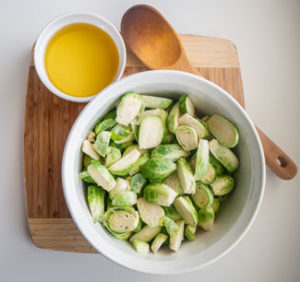 Brussels sprouts halved in a bowl alongside avocado oil and large wooden spoon